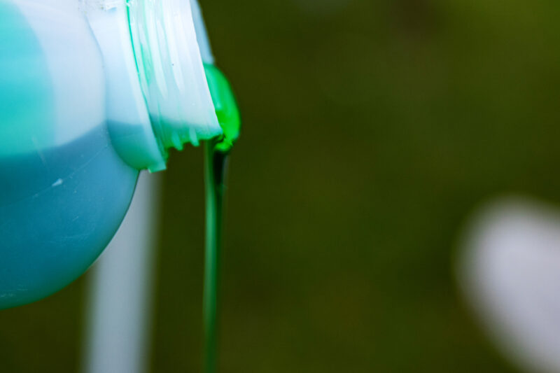 Closeup of vibrant green detergent pouring from a white plastic bottle against a blurred background. Green liquid flowing from a container. Copy space. Selective focus